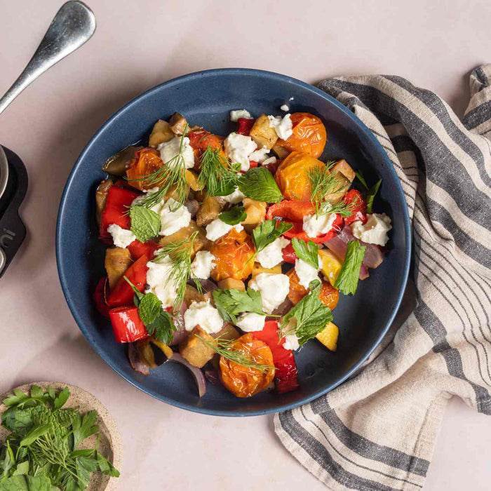 Courges, aubergines, oignons, tomates cerises et poivrons rôtis lentement dans une plat à rôtir Ooni, à côté d'un bol de légumes rôtis lentement avec de la feta et des herbes fraîches sur une table.