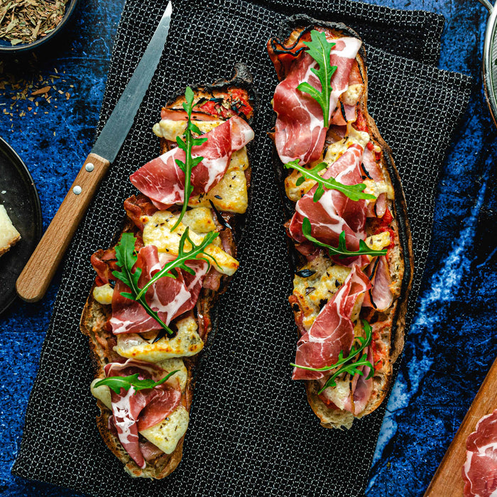 Cooked Corsican pizza baguette with Corsican cheese, pancetta, coppa, arugula and herbes de Provence on a table surrounded by a plate of cheese, bowl of arugula and serving board of pancetta. Emilia Sernagiotto