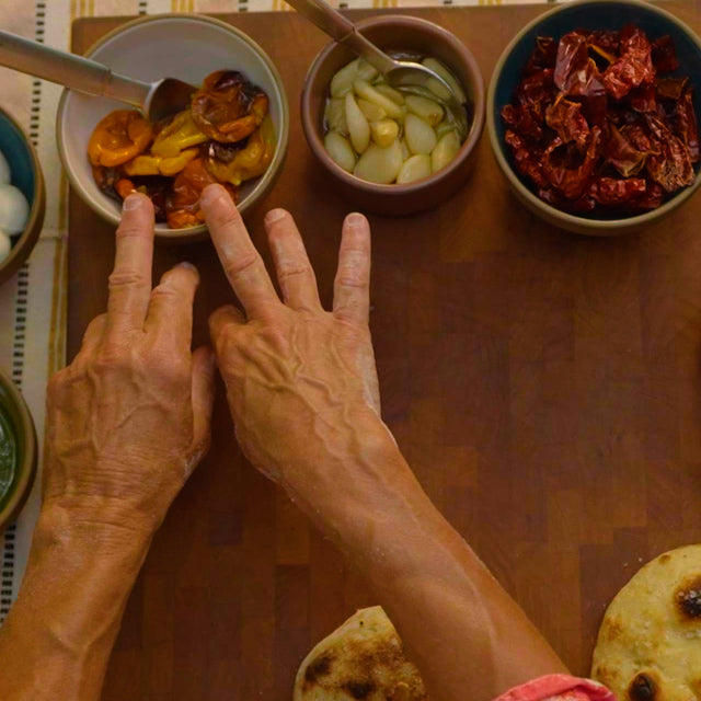 Deux mains qui touchent un bol de petits poivrons marinés à côté de bols de pesto au basilic, de boules de mozzarella, d'ail confit, de radis et de sel.