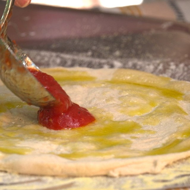 Ladel spooning tomato sauce on dough covered in olive oil on an Ooni Pizza Peel.