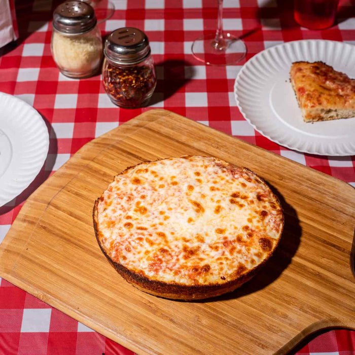 Pizza pan à la sauce tomate et à la mozarella sur une planche à découper en bambou Ooni sur une table avec des assiettes de tranches de pizza.